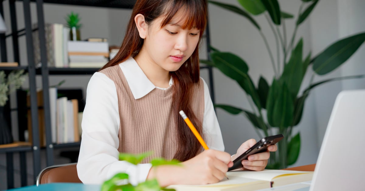 A female student studying at her laptop using her phone, notebook, and pencil.