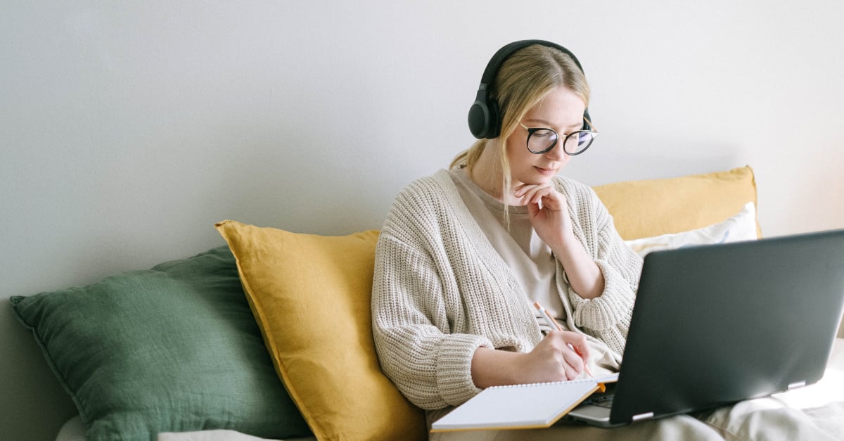 Student reclining against cushions, wearing headphones, and studying with a notepad and laptop.