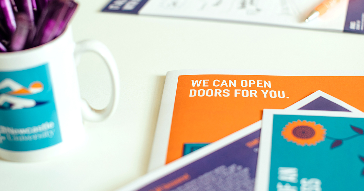 A white desk with mug and pamphlet out of focus in the background. A pile of pamphlets are in-focus in the foreground, featuring the words 'We can open doors for you'.