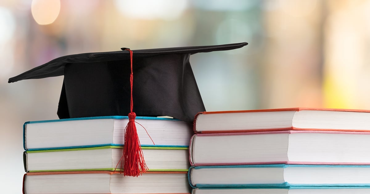 Two piles of books, topped with a black graduation cap with a red tassel.